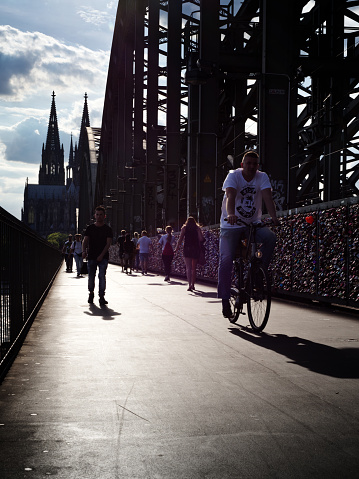 A man on a bicycle wearing a white shirt and pedestrians crossing Rhine river on Hohenzollern bridge (Hohenzollernbrücke) in Cologne with the famous landmark Cologne Cathedral in the background. Cologne, Germany. 05/16/2017