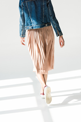 Rear view, low angle shot of woman in beige skirt and denim jacket walking in white studio