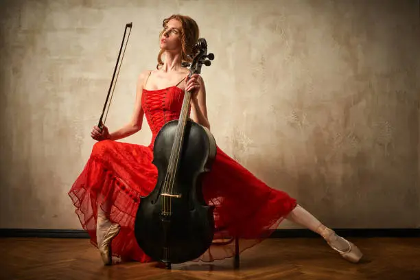 Female ballet dancer in red dress and pointe playing on antique black cello