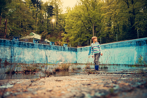 Little boy wearing galoshes is playing in a dirty old abadoned swimming pool. The boy is looking for frogs in a small pond on the base of swimming pool.