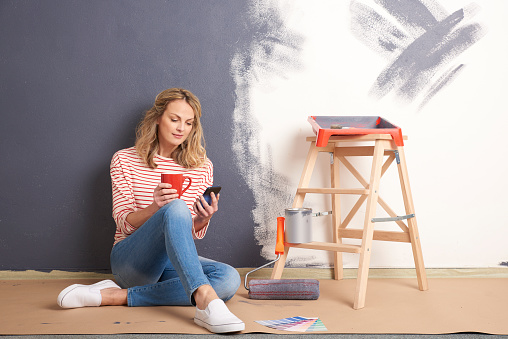 Shot of a smiling middle aged woman holding in hand a cup of tea and using her handy while painting wall in her new home under construction.