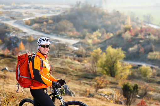 cyclist-traveler with a backpack on mountain bike rides on a mountain trail in spring season. countryside. beautiful landscape. cyclist in white helmet with red backpack.