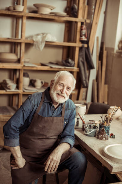 istock Senior potter in apron sitting at table and looking at camera at manufacturing 684987942