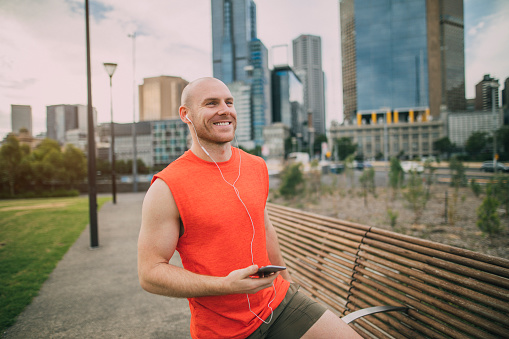 Mature man dressed in gym clothing using a smartphone with earphones plugged in, he is standing in a public park using his smartphone to prepare a music playlist for his run.