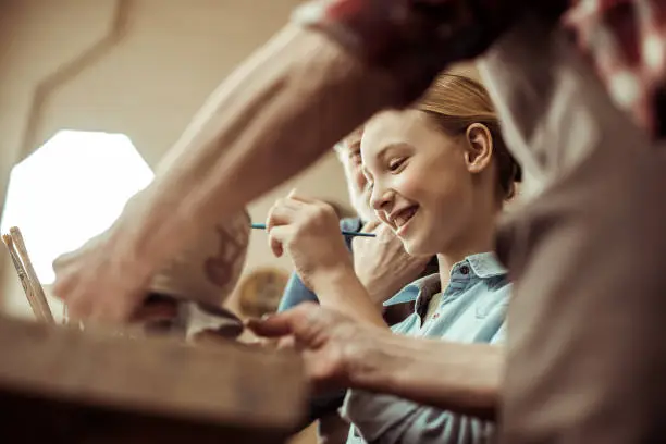 Photo of Close up of girl painting clay pot and grandparents helping at workshop