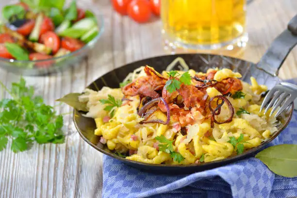 Swabian spaetzle with sauerkraut, fried bacon and onion rings on a wooden table with a Munich beer and a side salad