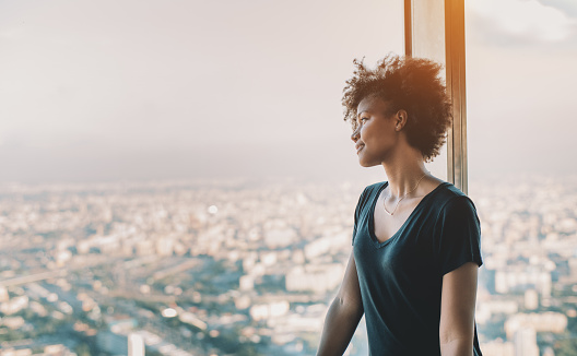 Attractive young mixed girl in black shirt and with curly african hair is standing near window of very high tower observation point and pensively looking on evening cityscape of modern metropolitan