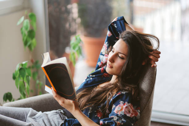 Young woman reading at home Vintage toned portrait of a young woman relaxing and reading after work, in the bright and beautiful apartment on the high floor in Belgrade, Serbia. She is wearing casual, everyday clothes. tired after work stock pictures, royalty-free photos & images