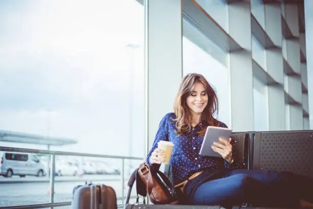 Shot of happy young woman with coffee waiting her flight at airport lounge and reading ebook on her digital tablet.