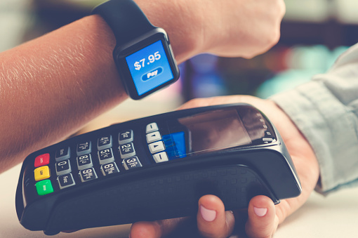 Paying with a smart watch. They are holding it close to a credit card reader. The screen says $7.95 with the word pay. The shopkeeper is holding the credit card reader