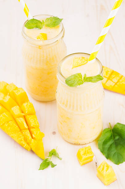 istock Freshly blended yellow mango fruit smoothie in glass jars with straw, mint leaves, mango slices, close up. Soft white wooden board background. 685430796