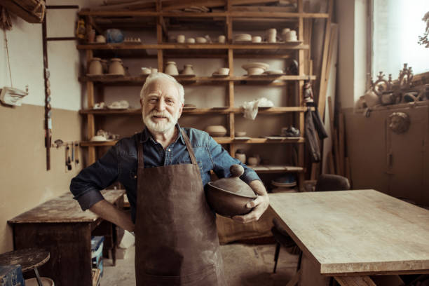istock Front view of proud senior potter in apron standing with ceramic bowl at workshop 685464830