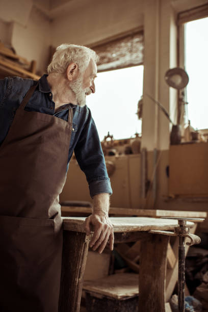 istock Side view of senior potter in apron standing and leaning on table at workshop 685489094