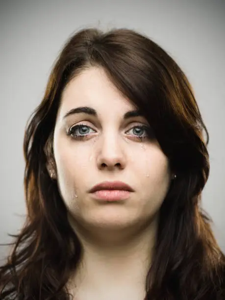Close-up portrait of sad young caucasian woman. Crying female against gray background with tears in her face. Vertical studio photography from a DSLR camera. Sharp focus on eyes.