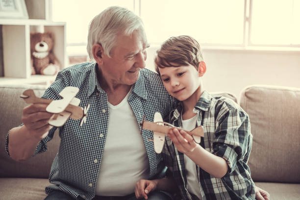 Grandpa and grandson are playing with toy planes and smiling while sitting on couch at home