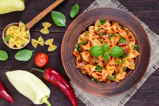 Pasta farfalle with chicken, tomato sauce and basil in a clay bowl on dark wooden background. The top view