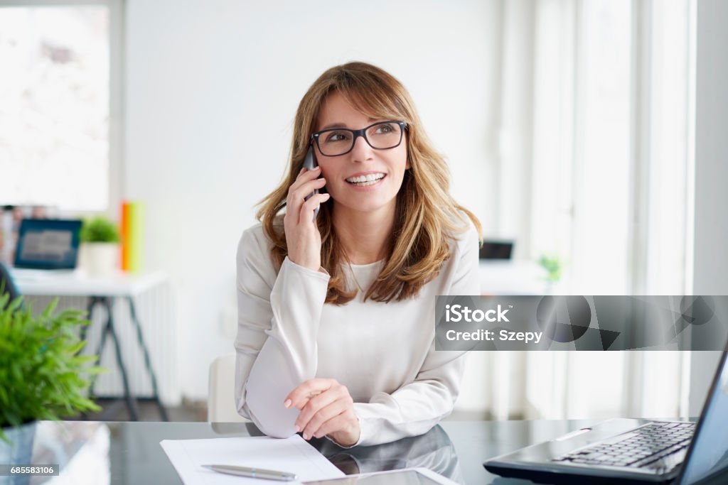 Business conversation Shot of a beautiful mature businesswoman talking with somebody while sitting at office desk and working on laptop. Women Stock Photo