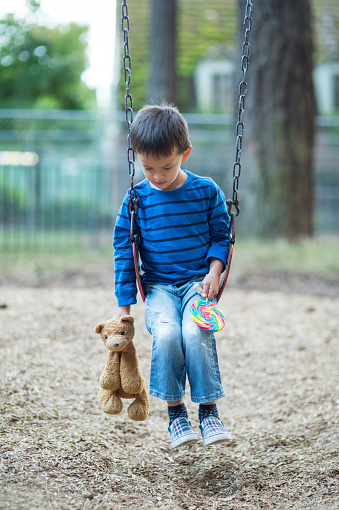 Young boy swinging by himself on playground holding teddy bear and lollipop.