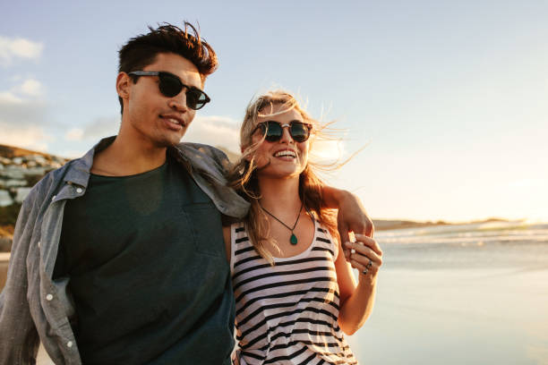 Young couple enjoying a summer day on seashore Portrait of handsome young man with his beautiful girlfriend on beach. Young couple enjoying a summer day on seashore. beach fashion stock pictures, royalty-free photos & images