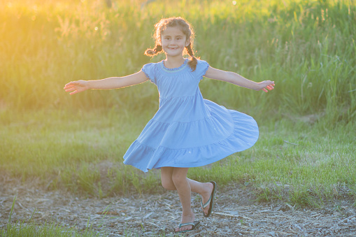 Little girl is dancing and spinning around outside wearing a blue dress and pigtails as she is backlit by a summer sunset.