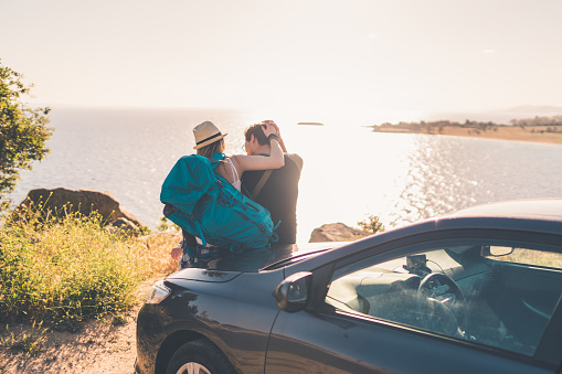 Couple sitting on the car hood with backpacks and bags on and looking at the beautiful sea view and enjoying time together