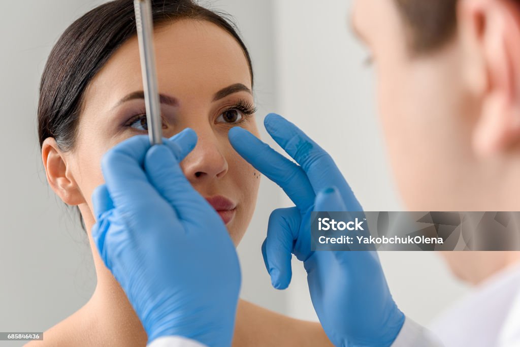 Physician examining face of serene female Focus on doctor hands showing where correct nose of calm woman. She watching at therapeutic Nose Stock Photo