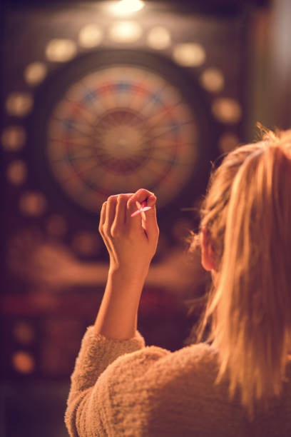 Rear view of a woman playing darts in entertainment club. Back view of woman aiming at dartboard in a pub. Focus is on her hand. woman darts stock pictures, royalty-free photos & images