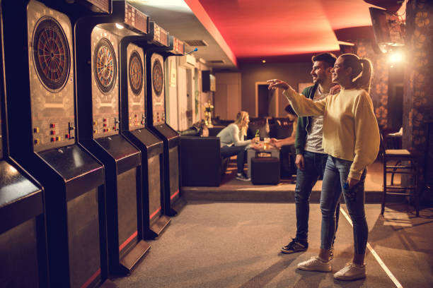 Young happy couple playing darts in entertainment club. Full length of smiling couple enjoying in playing darts in a pub. There are people in the background. woman darts stock pictures, royalty-free photos & images