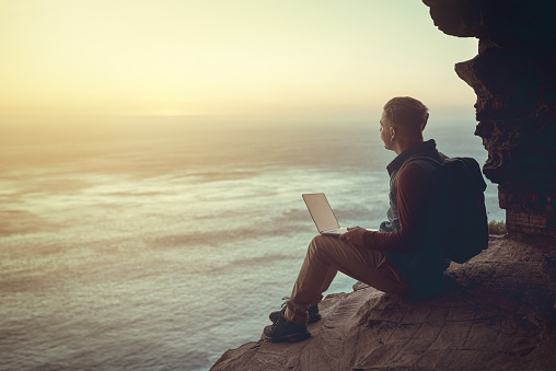 Full length shot of a young man using his laptop while sitting on a cliff in the mountains