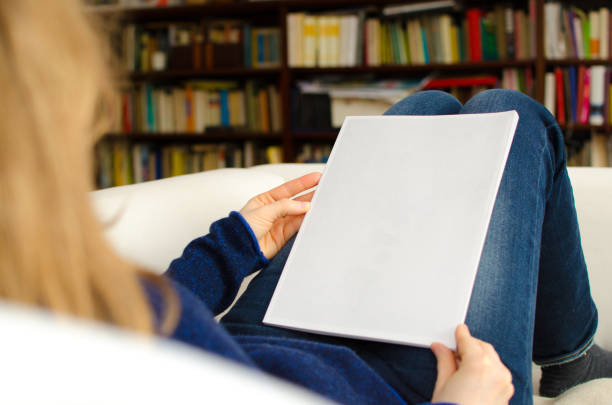 blond woman sitting at home on a sofa and is holding a blank brochure in her hands - newspaper reading blank women imagens e fotografias de stock