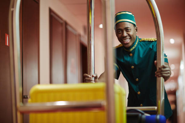 Smiling Bellboy Carrying Luggage in Cart Portrait of smiling African bellhop helping guests, pushing luggage cart delivering bags to hotel rooms bellhop stock pictures, royalty-free photos & images