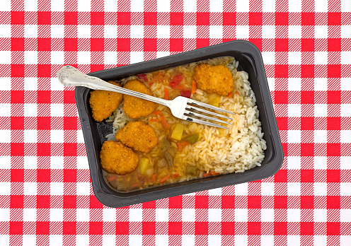 Top view of a TV dinner of sweet and sour chicken in an black plastic tray with a fork in the food on a bright red and white tablecloth.