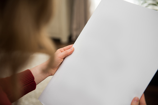 A woman is holding a blank brochure in her hands and is looking at the white page of the magazine. The document is left blank for your own designs or pictures. The photo was taken out of a personal perspektive in a domestic living room.