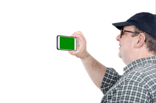 Fat man with chubby face shoots with mobile phone. Fifty years old man looks at chroma key screen. He wears plaid shirt and black cap. Tourism concept. Horizontal mid shot on white background