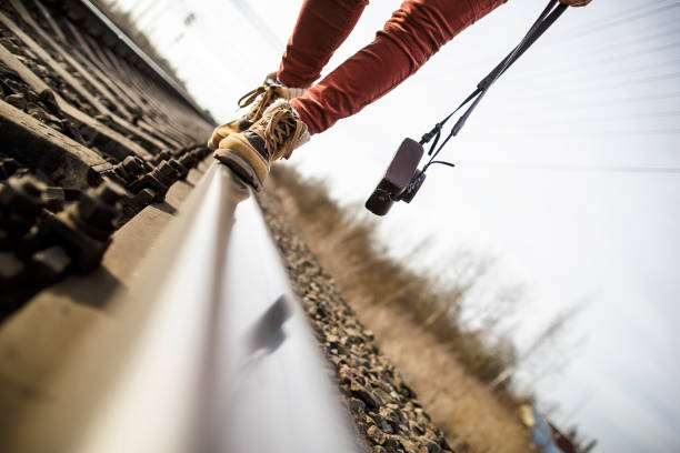 Young woman with legs, dressed in boots walking on railroad tracks, holding a camera in her hands