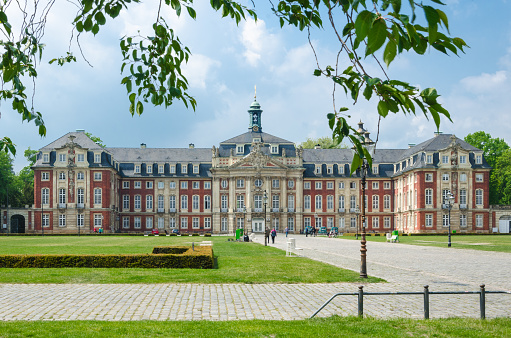 Münster, Germany - May 17, 2016: The background photo shows the university building and campus of the University of Münster (Westfälische Wilhelms-Universität Münster, WWU). The name of the building is Fürstbischöfliches Schloss Münster.  There are some people walking by the building. The photo was taken on a sunny summer's day.
