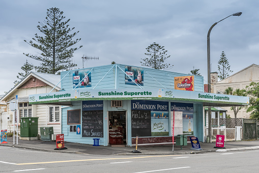 Napier, New Zealand - March 9, 2017: Sunshine Superette is a corner store selling groceries, newspapers and basic household products. Light blue paint, colored advertisement, cloudy sky and street scene.