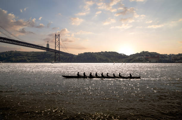 日没時に吊り橋を背景に海のボートに乗っている人々のシルエット。リスボン、ポルトガル。 - 漕ぐ ストックフォトと画像