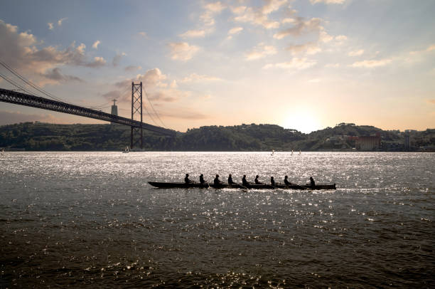 silhueta de pessoas em barco de remo no mar com ponte suspensa ao fundo ao pôr do sol. lisboa, portugal. - remo desportivo - fotografias e filmes do acervo