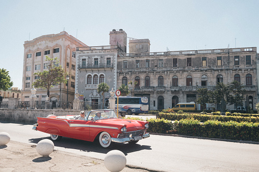 This is a horizontal, color photograph of two men driving in a vintage, red, Buick convertible on the streets of travel destination Havana, Cuba. Photographed on a sunny day. CreativeContentBrief 696189239