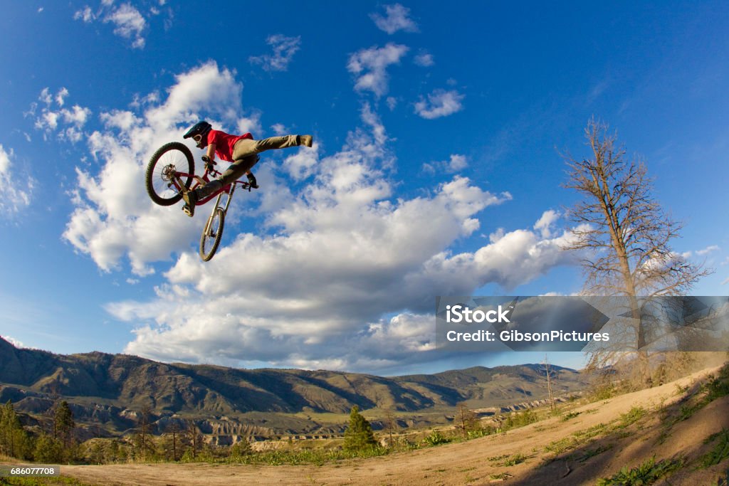 A young man does a nac nac trick on his downhill-style mountain bike. He takes one foot off the pedal and pushes the bike sideways and forward with the other foot. He wears jeans, a t-shirt and a full face bicycle helmet. Adult Stock Photo