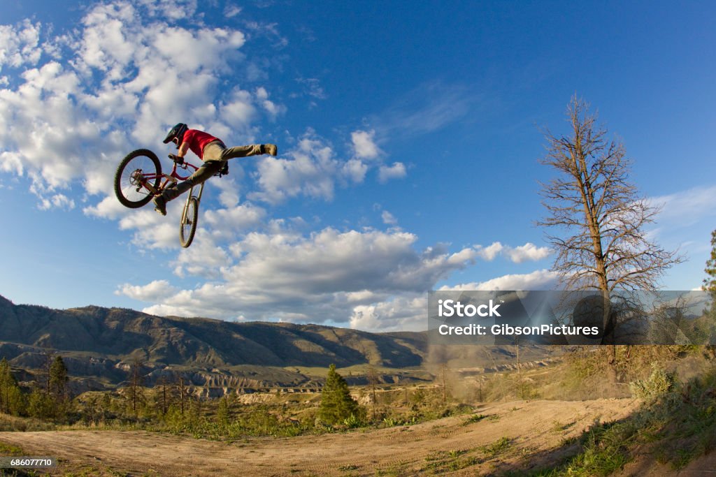 A young man does a nac nac trick on his downhill-style mountain bike. He takes one foot off the pedal and pushes the bike sideways and forward with the other foot. He wears jeans, a t-shirt and a full face bicycle helmet. Active Lifestyle Stock Photo