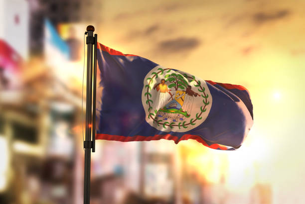 флаг белиза против размытого фона города на восходе подсветки - flag of belize стоковые фото и изображения