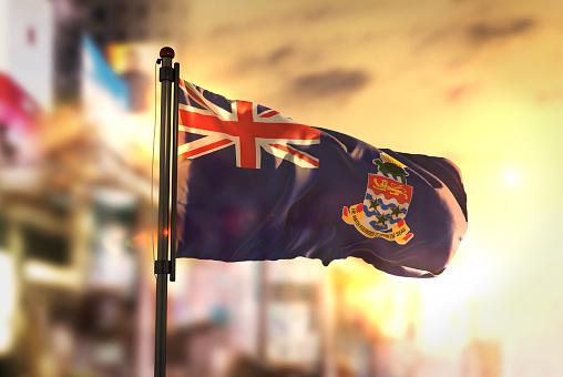 Cayman Islands Flag Against City Blurred Background At Sunrise Backlight