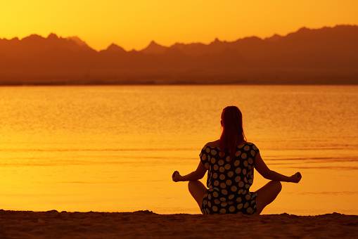 rear view of young woman relaxing and sitting in lotus position on the beach, enjoying the sunset and the silence.