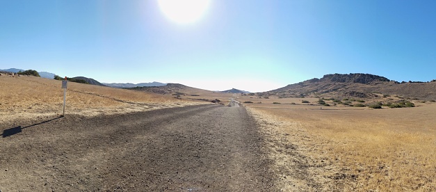Panoramic dry landscape with bright sun, blue sky, and dirt trail.