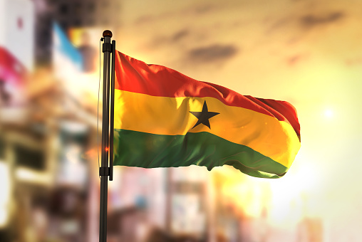 Ghana Flag Against City Blurred Background At Sunrise Backlight