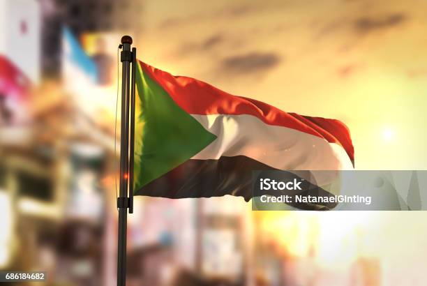 Sudan Flag Against City Blurred Background At Sunrise Backlight Stock Photo - Download Image Now