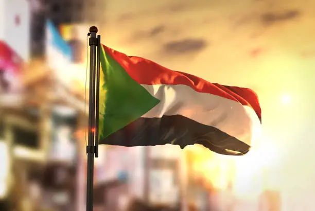 Photo of Sudan Flag Against City Blurred Background At Sunrise Backlight