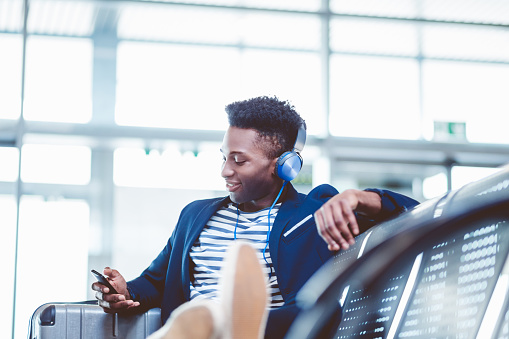 Young african man wearing headphones and listening to music from mobile phone while waiting for flight at airport departure area.